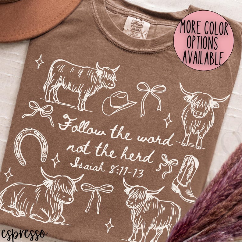 May include: Espresso-colored t-shirt with a white graphic design featuring Highland cows, a cowboy hat, bows, a horseshoe, and a cowboy boot. The text "Follow the word not the herd Isaiah 8:11-13" is also included. A pink circle indicates more color options.