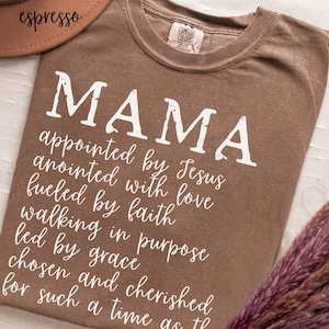 May include: Espresso-colored t-shirt with the word "MAMA" in large white letters. Below, a script lists inspirational phrases. The shirt is a Comfort Colors brand.