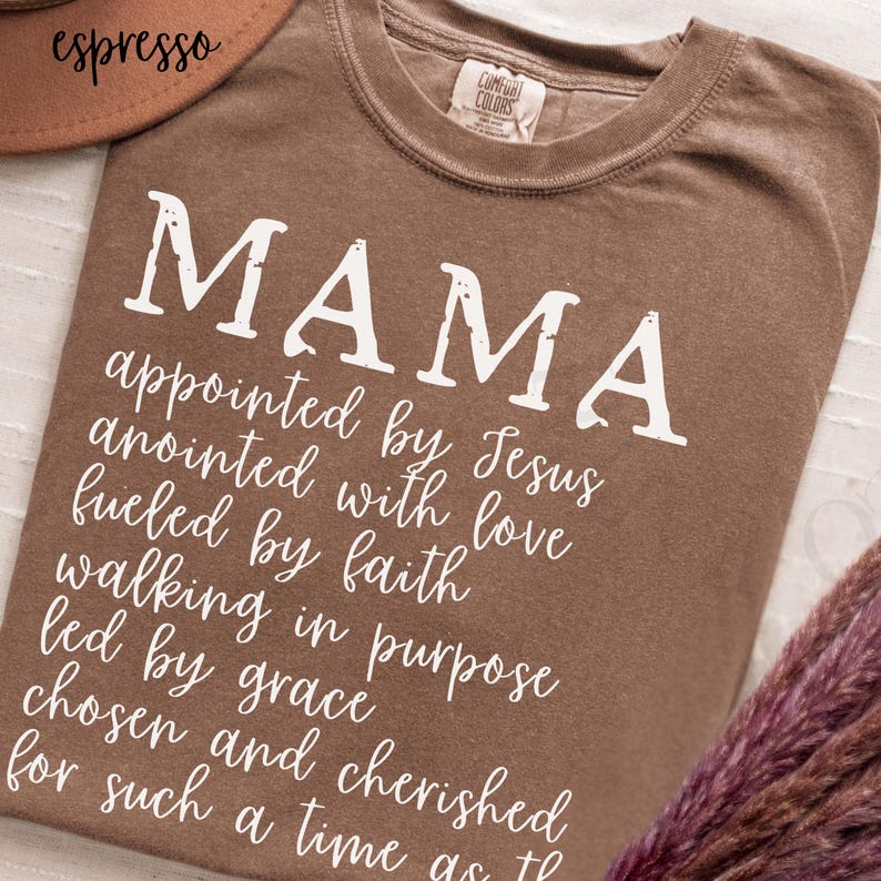May include: Espresso-colored t-shirt with the word "MAMA" in large white letters. Below, a script lists inspirational phrases. The shirt is a Comfort Colors brand.