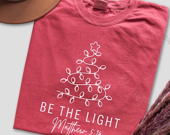 May include: Crimson red t-shirt with a white Christmas tree design made of string lights. The text "BE THE LIGHT Matthew 5:14" is printed below the tree. A brown hat is visible in the upper left corner.