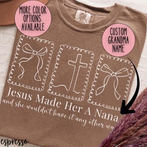 Pu&ograve; includere: T-shirt color espresso con grafica bianca di una croce e fiocchi. Il testo recita "Jesus Made Her A Nana and she wouldn't have it any other wa." Cerchi rosa dicono "More Color Options Available" e "Custom Grandma Name."