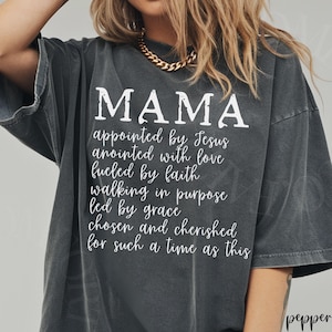 May include: Dark gray t-shirt with the word "MAMA" in large white letters. Below, a list of inspirational phrases is printed in white. The shirt has a relaxed fit and a vintage look.