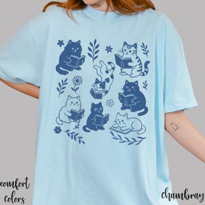 以下が含まれることがあります： 本を読む猫のネイビーブルーのイラストが描かれた水色のTシャツ。花と葉のモチーフが周りに描かれています。「comfort colors」の文字が左下に、「chambray」の文字が右下にプリントされています。