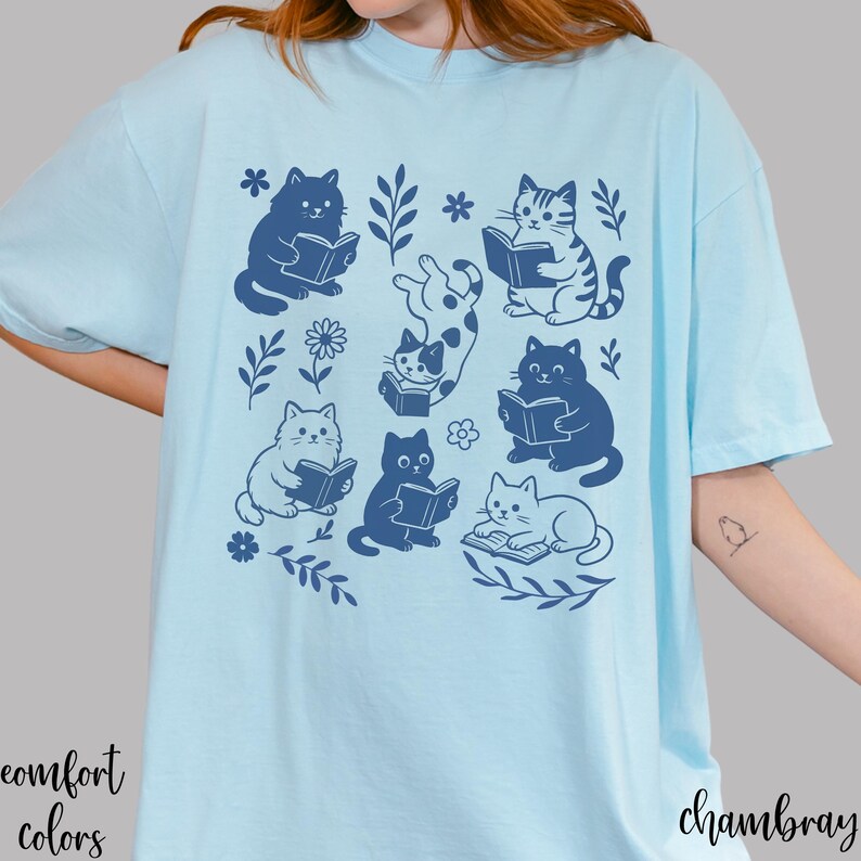 以下が含まれることがあります： 本を読む猫のネイビーブルーのイラストが描かれた水色のTシャツ。花と葉のモチーフが周りに描かれています。「comfort colors」の文字が左下に、「chambray」の文字が右下にプリントされています。