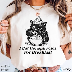 May include: Ivory-colored t-shirt featuring a black and white graphic of a cat wearing a party hat and eating from a bowl. The text "1 Eat Conspiracies For Breakfast" is below the graphic. The shirt is labeled "comfort colors".