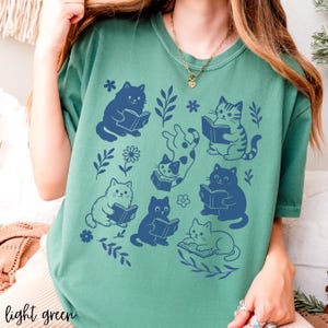 以下が含まれることがあります： 本を読む猫の青いグラフィックが特徴の、明るい緑色のTシャツ。花柄と葉柄のデザインが施されています。「light green」の文字がシャツの下部にプリントされています。
