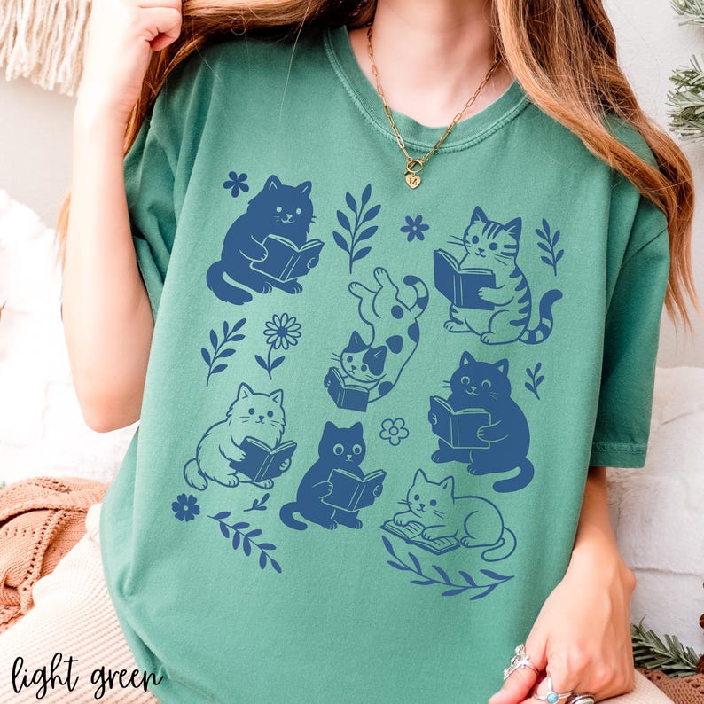 以下が含まれることがあります： 本を読む猫の青いグラフィックが特徴の、明るい緑色のTシャツ。花柄と葉柄のデザインが施されています。「light green」の文字がシャツの下部にプリントされています。