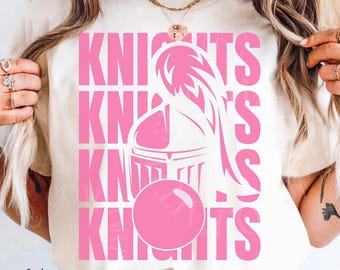 May include: Ivory-colored t-shirt with the word "KNIGHTS" in pink, repeated vertically. A pink knight's helmet and a pink sphere are also featured. The words "comfort colors" are printed in the bottom left corner.