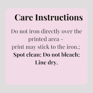 May include: A light pink rectangular card with rounded corners displays care instructions in black text. The instructions advise against ironing directly over the printed area, spot cleaning, avoiding bleach, and line drying.