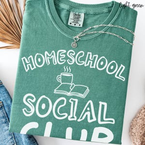 Op de afbeelding: Een lichtgroen T-shirt met korte mouwen en witte opdruk met de tekst HOMESCHOOL SOCIAL CLUB, inclusief een afbeelding van een dampende koffiemok en een open boek. Een zilverkleurige ketting met een ronde hanger rust op de kraag van het shirt.