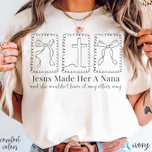 Pu&ograve; includere: T-shirt color avorio con la scritta "Jesus Made Her A Nana and she wouldn't have it any other way." La maglietta presenta tre disegni a tratto nero: due fiocchi e una croce, ciascuno all'interno di una cornice decorativa. Sono visibili anche le parole "comfort colors".