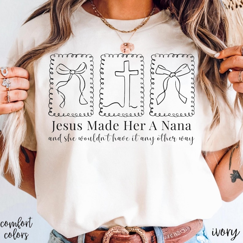 Pu&ograve; includere: T-shirt color avorio con la scritta "Jesus Made Her A Nana and she wouldn't have it any other way." La maglietta presenta tre disegni a tratto nero: due fiocchi e una croce, ciascuno all'interno di una cornice decorativa. Sono visibili anche le parole "comfort colors".