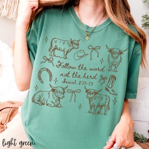 May include: Light green t-shirt with a brown graphic design featuring Highland cows, a cowboy hat, a horseshoe, bows, and a cowboy boot. The text "Follow the word not the herd" and Bible verse Isaiah 8:11-13 are also printed on the shirt.