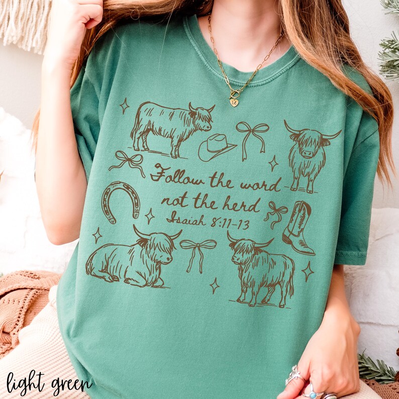 May include: Light green t-shirt with a brown graphic design featuring Highland cows, a cowboy hat, a horseshoe, bows, and a cowboy boot. The text "Follow the word not the herd" and Bible verse Isaiah 8:11-13 are also printed on the shirt.