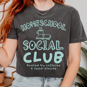 Op de afbeelding: Een antracietgrijs T-shirt met korte mouwen en de lichtblauwe tekst "HOMESCHOOL SOCIAL CLUB" boven een afbeelding van een dampende koffiemok en een open boek. Onder de afbeelding staat de tekst "Fueled by caffeine & read-alouds" in het lichtblauw.