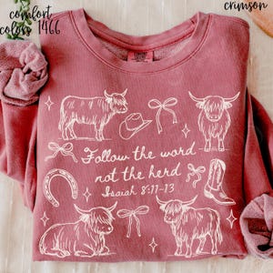 May include: Crimson-colored sweatshirt with white graphic of Highland cows, cowboy hat, horseshoe, bows, and a cowboy boot. Text reads "Follow the word not the herd Isaiah 8:11-13". The sweatshirt is Comfort Colors brand.
