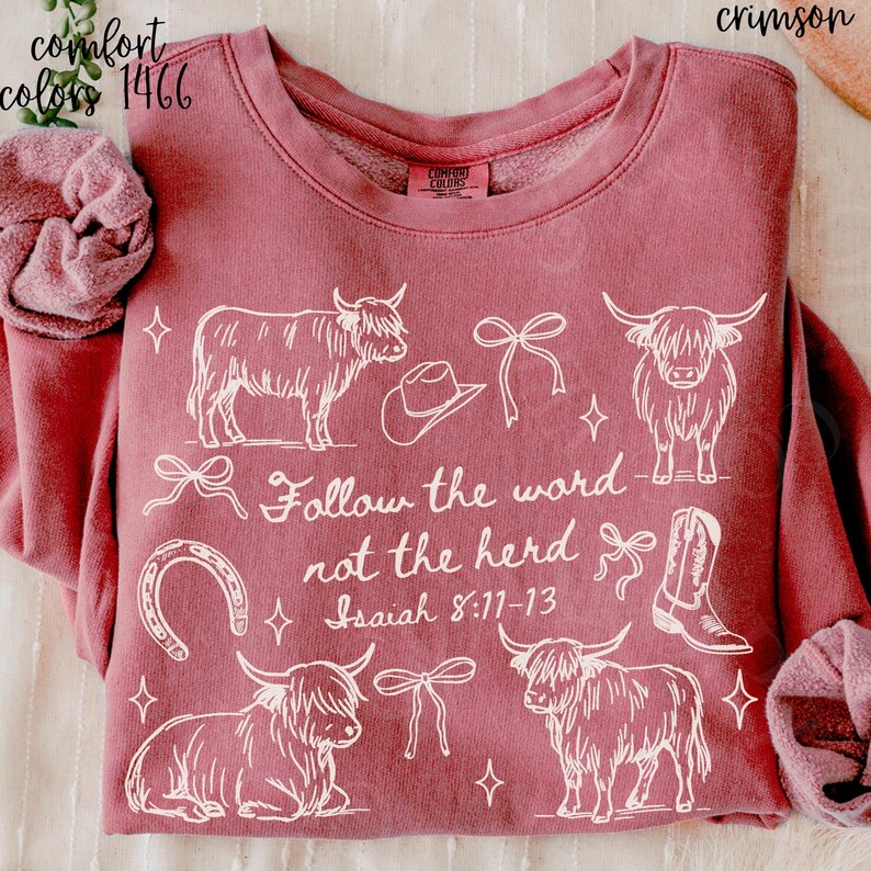 May include: Crimson-colored sweatshirt with white graphic of Highland cows, cowboy hat, horseshoe, bows, and a cowboy boot. Text reads "Follow the word not the herd Isaiah 8:11-13". The sweatshirt is Comfort Colors brand.