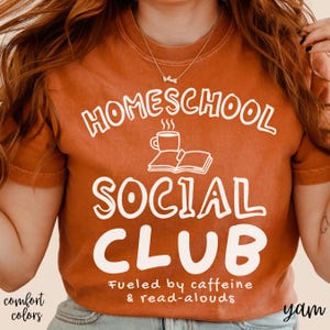 Op de afbeelding: Een kort mouwloos t-shirt in gebrand oranje met witte tekst HOMESCHOOL SOCIAL CLUB, een afbeelding van een dampende koffiemok en een open boek, plus de tekst Fueled by caffeine & read-alouds in een kleiner wit lettertype.