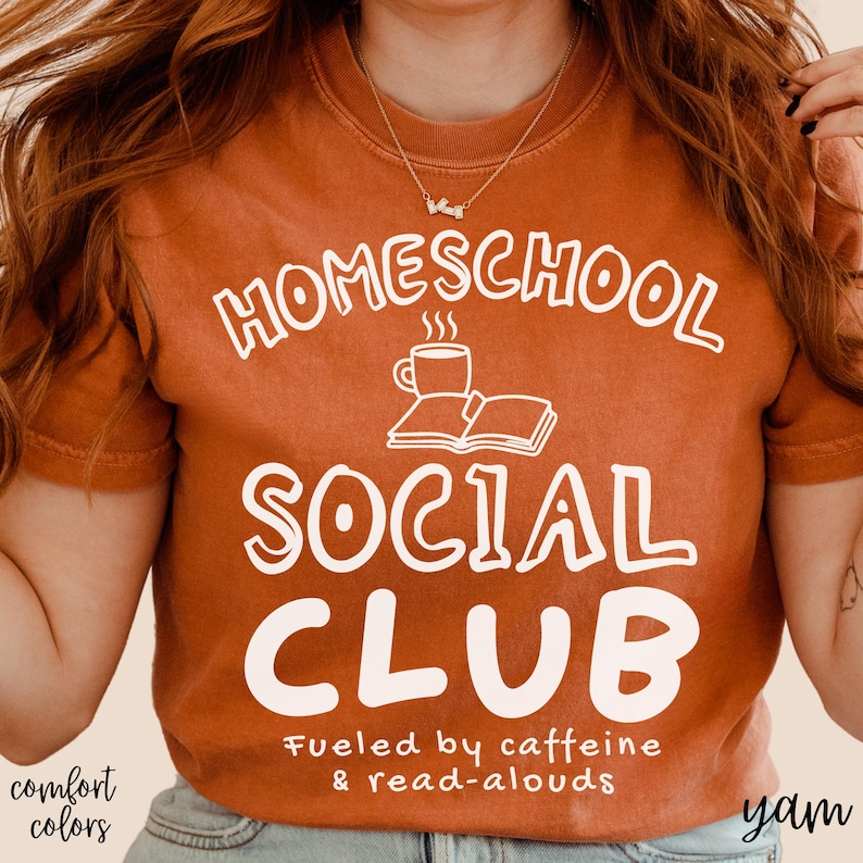 Op de afbeelding: Een kort mouwloos t-shirt in gebrand oranje met witte tekst HOMESCHOOL SOCIAL CLUB, een afbeelding van een dampende koffiemok en een open boek, plus de tekst Fueled by caffeine & read-alouds in een kleiner wit lettertype.