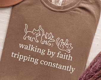 May include: Espresso-colored t-shirt with a white graphic of three cats and the text "walking by faith tripping constantly." The shirt has a crew neck and a Comfort Colors tag.