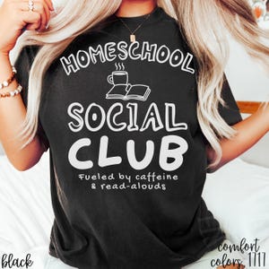 Op de afbeelding: Een zwart t-shirt met witte tekst die HOMESCHOOL SOCIAL CLUB leest, met een afbeelding van een dampende koffiekop en een open boek erboven. Onder de hoofdtekst staat in een kleiner lettertype Fueled by caffeine & read-alouds.