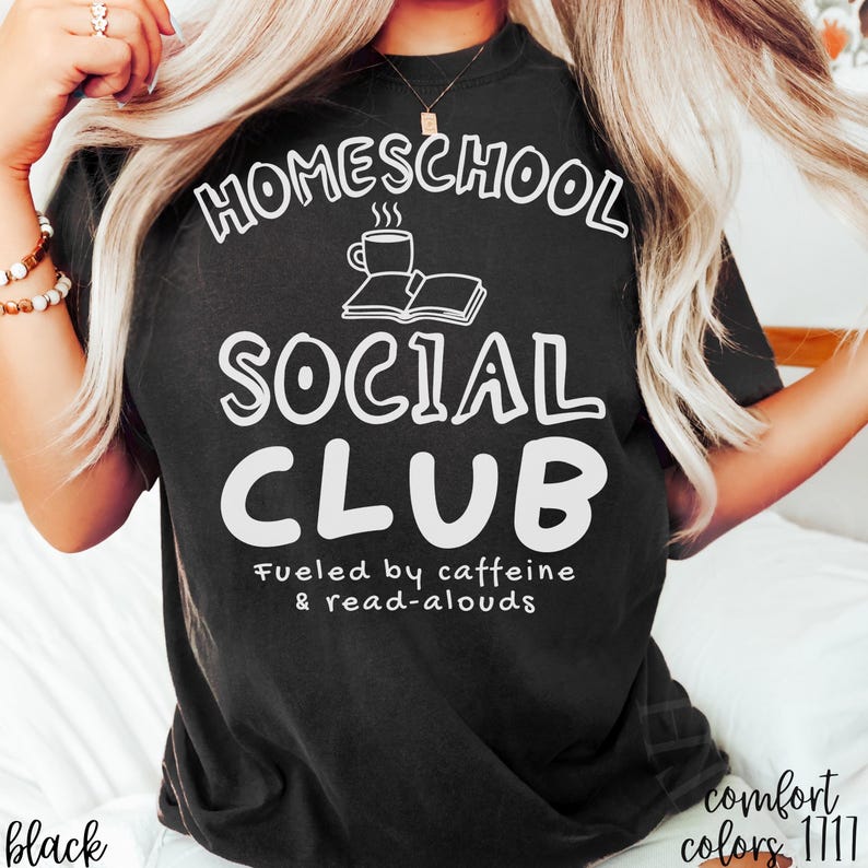 Op de afbeelding: Een zwart t-shirt met witte tekst die HOMESCHOOL SOCIAL CLUB leest, met een afbeelding van een dampende koffiekop en een open boek erboven. Onder de hoofdtekst staat in een kleiner lettertype Fueled by caffeine & read-alouds.