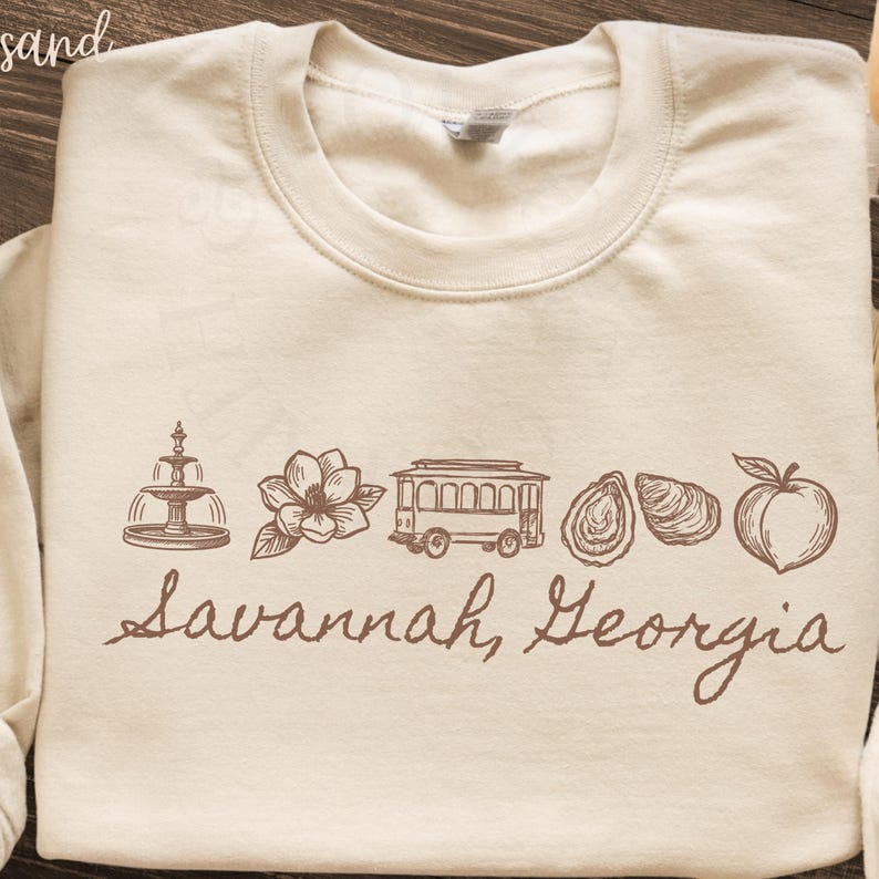 Puede incluir: Sudadera color crema con un dise&ntilde;o gr&aacute;fico marr&oacute;n que presenta una fuente, una flor, un tranv&iacute;a, ostras y un melocot&oacute;n. Las palabras "Savannah, Georgia" est&aacute;n escritas en cursiva debajo.