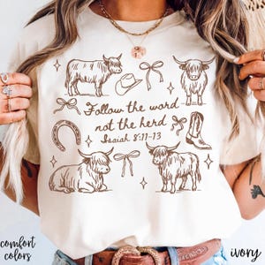 May include: Ivory-colored t-shirt featuring brown illustrations of Highland cows, a cowboy hat, bows, a horseshoe, and a cowboy boot. The text "Follow the word not the herd Isaiah 8:11-13" is also printed on the shirt. The words "comfort colors" are printed on the bottom left.
