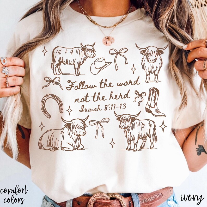 May include: Ivory-colored t-shirt featuring brown illustrations of Highland cows, a cowboy hat, bows, a horseshoe, and a cowboy boot. The text "Follow the word not the herd Isaiah 8:11-13" is also printed on the shirt. The words "comfort colors" are printed on the bottom left.