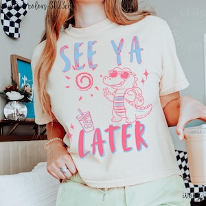 May include: Ivory-colored t-shirt with the text "SEE YA LATER" in blue and pink. A cartoon alligator wearing sunglasses and a backpack is featured. The shirt also has a graphic of a drink and a sun.