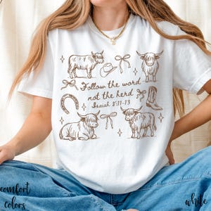 May include: White t-shirt with a brown graphic design featuring Highland cows, a cowboy hat, boots, and bows. The text "Follow the word not the herd Isaiah 8:11-13" is also printed on the shirt.