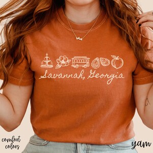 Puede incluir: Camiseta color &oacute;xido con un gr&aacute;fico blanco de los monumentos de Savannah, Georgia, incluyendo una fuente, una flor, un tranv&iacute;a, ostras y un melocot&oacute;n. El texto "Savannah, Georgia" est&aacute; escrito debajo.