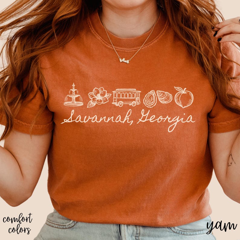 Puede incluir: Camiseta color &oacute;xido con un gr&aacute;fico blanco de los monumentos de Savannah, Georgia, incluyendo una fuente, una flor, un tranv&iacute;a, ostras y un melocot&oacute;n. El texto "Savannah, Georgia" est&aacute; escrito debajo.