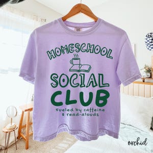 Op de afbeelding: Een lichtpaars t-shirt met korte mouwen en donkergroene tekst "HOMESCHOOL SOCIAL CLUB" met een afbeelding van een koffiekopje en een open boek. Onder de hoofdtekst staat in het groen de tekst "Fueled by caffeine & read-alouds" op het shirt.