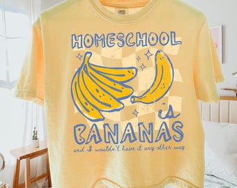 May include: A pale yellow short-sleeve t-shirt featuring a graphic of yellow bananas and a checkered background. The text reads: HOMESCHOOL is BANANAS and I wouldn't have it any other way. The shirt is labelled as a boxy tee, Comfort Colors 3023cl, butter.
