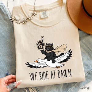 May include: Ivory-colored Comfort Colors t-shirt featuring a cartoon panther wearing sunglasses and a scarf, riding a goose. The panther is holding a foam finger. The text "WE RIDE AT DAWN" is printed below the graphic.