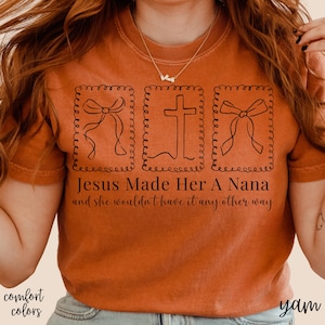 Pu&ograve; includere: T-shirt color arancione bruciato con la scritta "Jesus Made Her A Nana and she wouldn't have it any other way." Il design include tre immagini incorniciate in nero di un fiocco e una croce. Le parole "comfort colors" sono in basso a sinistra.