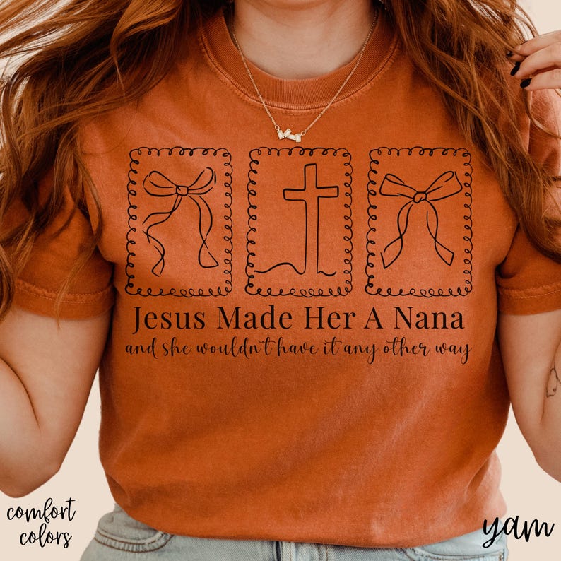 Pu&ograve; includere: T-shirt color arancione bruciato con la scritta "Jesus Made Her A Nana and she wouldn't have it any other way." Il design include tre immagini incorniciate in nero di un fiocco e una croce. Le parole "comfort colors" sono in basso a sinistra.