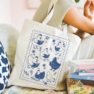 May include: A cream-coloured tote bag with a blue stamp design. The design features illustrations of chickens reading books, with floral and leaf accents. The bag has two handles and is made of a sturdy fabric, creating a whimsical and literary theme.