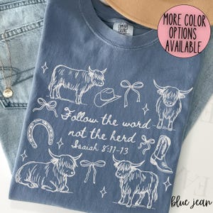 May include: Blue jean colored t-shirt with a white graphic design featuring Highland cows, a cowboy hat, bows, a horseshoe, and a cowboy boot. The text "Follow the word not the herd Isaiah 8:11-13" is also printed on the shirt.