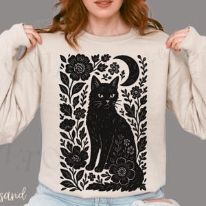 May include: Beige sweatshirt featuring a black cat illustration surrounded by flowers and a crescent moon. The design is a detailed, black and white graphic. The sweatshirt is a neutral color, suitable for casual wear.
