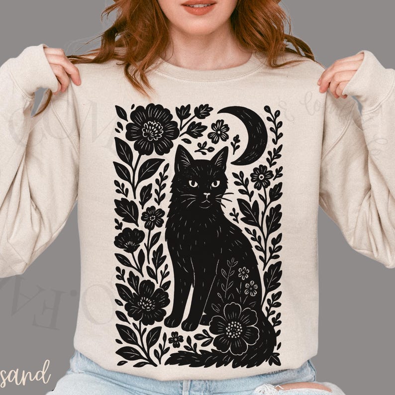 May include: Beige sweatshirt featuring a black cat illustration surrounded by flowers and a crescent moon. The design is a detailed, black and white graphic. The sweatshirt is a neutral color, suitable for casual wear.