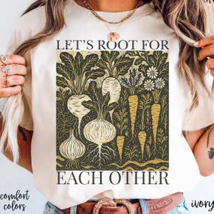 May include: Ivory-colored t-shirt featuring a botanical design with the text "LET'S ROOT FOR EACH OTHER." The graphic includes illustrations of root vegetables and flowers in shades of gold and black. The shirt is part of the "comfort colors" collection.