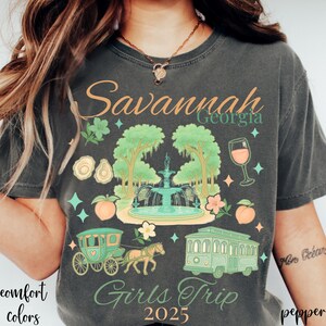 May include: Dark gray t-shirt with a colorful graphic design featuring the text "Savannah Georgia Girls Trip 2025." The design includes a fountain, trees, a horse-drawn carriage, a trolley, and peaches.