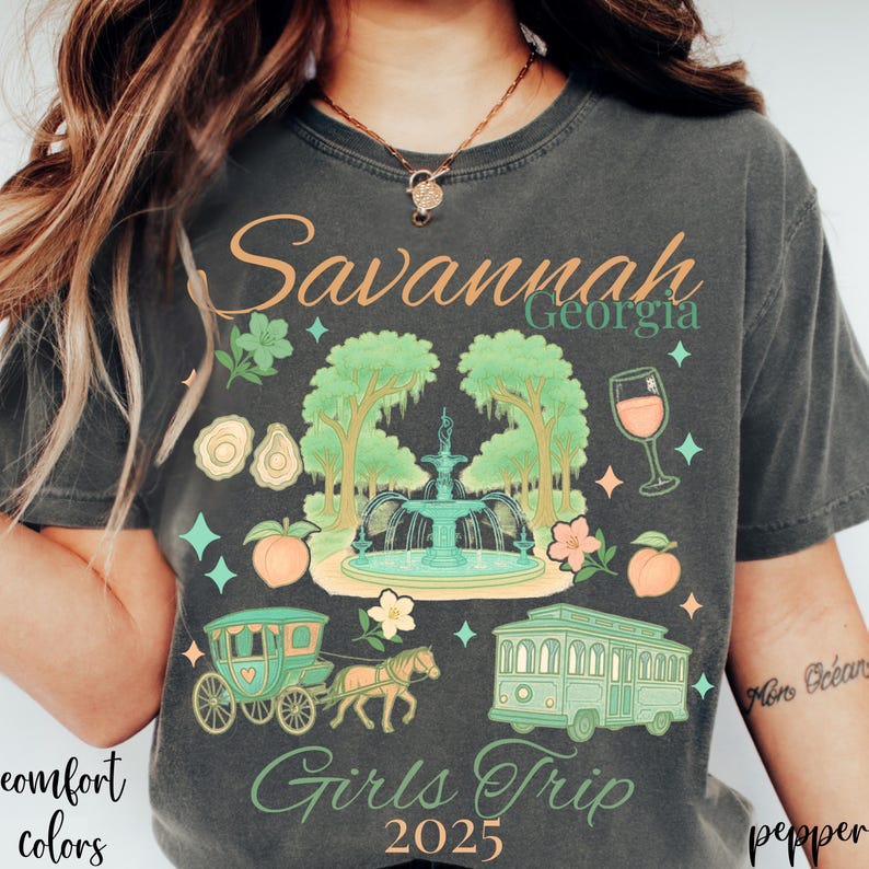 May include: Dark gray t-shirt with a colorful graphic design featuring the text "Savannah Georgia Girls Trip 2025." The design includes a fountain, trees, a horse-drawn carriage, a trolley, and peaches.