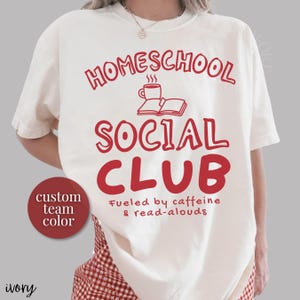 Op de afbeelding: Een ivoorkleurig t-shirt met rode tekst die HOMESCHOOL SOCIAL CLUB leest, met een afbeelding van een koffiemok en een open boek. Onder de tekst staat Fueled by caffeine & read-alouds. Een rode ronde badge bevat de tekst custom team color.
