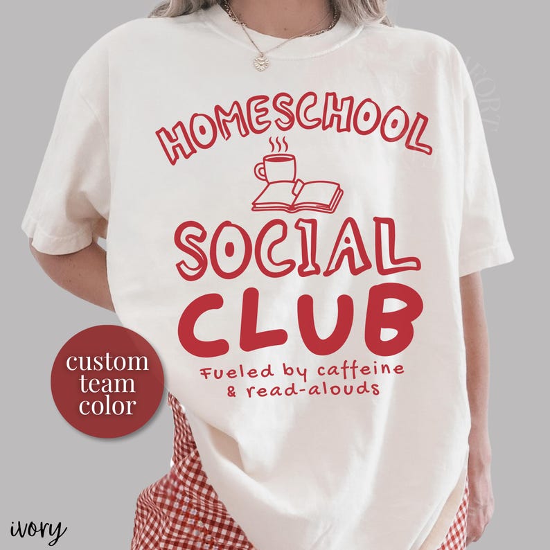 Op de afbeelding: Een ivoorkleurig t-shirt met rode tekst die HOMESCHOOL SOCIAL CLUB leest, met een afbeelding van een koffiemok en een open boek. Onder de tekst staat Fueled by caffeine & read-alouds. Een rode ronde badge bevat de tekst custom team color.