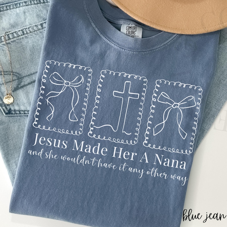 Pu&ograve; includere: T-shirt Comfort Colors blu jeans con testo e grafica bianchi. Il testo recita "Jesus Made Her A Nana and she wouldn't have it any other way." La grafica include una croce e due fiocchi.