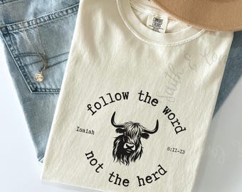 May include: Ivory-colored t-shirt with the text "follow the word not the herd" surrounding a black and white Highland cow graphic. The shirt also includes the text "Isaiah 8:11-13". A tan hat and blue jeans are in the background.