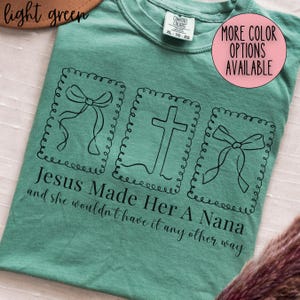 Pu&ograve; includere: T-shirt verde chiaro con la scritta "Jesus Made Her A Nana and she wouldn't have it any other way." Il design include tre illustrazioni incorniciate: due fiocchi e una croce. La maglietta ha un girocollo e un'etichetta "Comfort Colors".